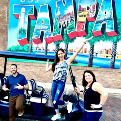 Three people posing cheerfully in front of a large, colorful "Tampa" mural. They are standing beside a golf cart.