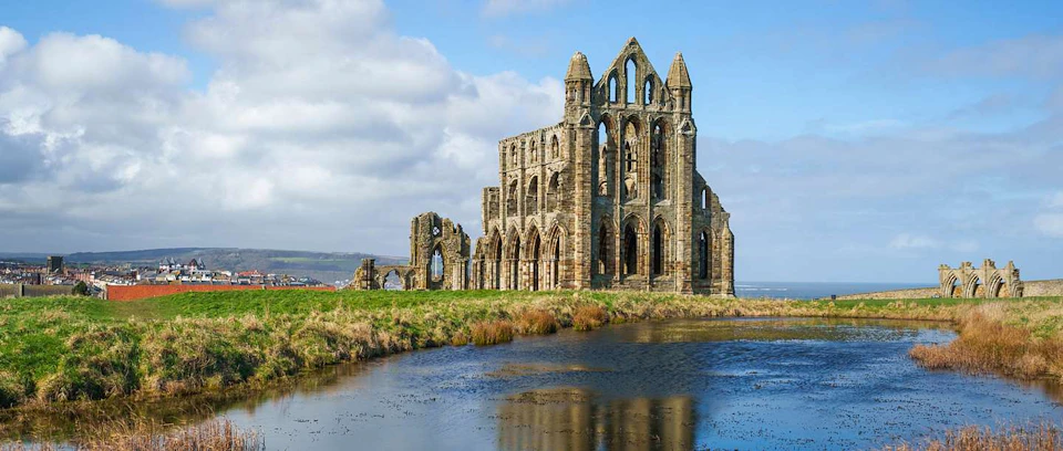 A large stone abbey ruin on a grassy hill overlooking a body of water, under a blue sky with scattered clouds.