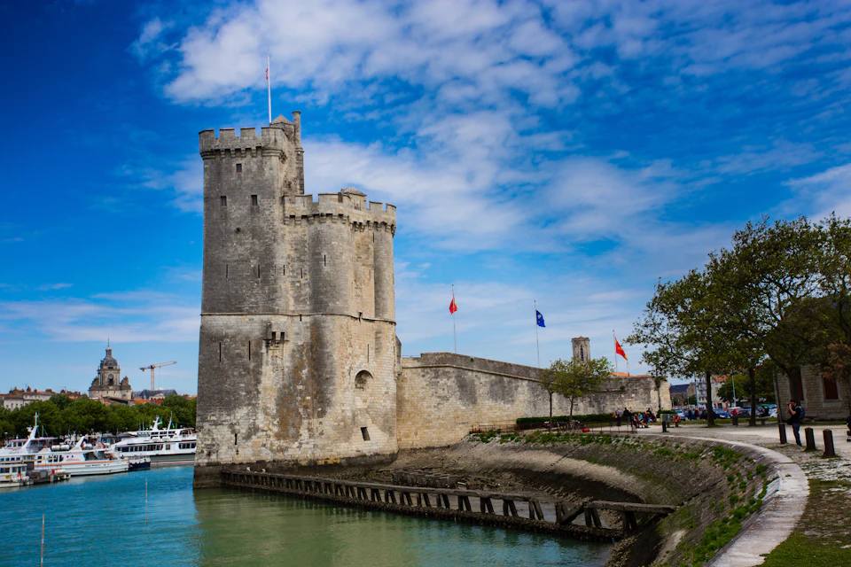 Historic stone fortress with flags and crenelated towers beside calm greenish water under a partly cloudy sky.