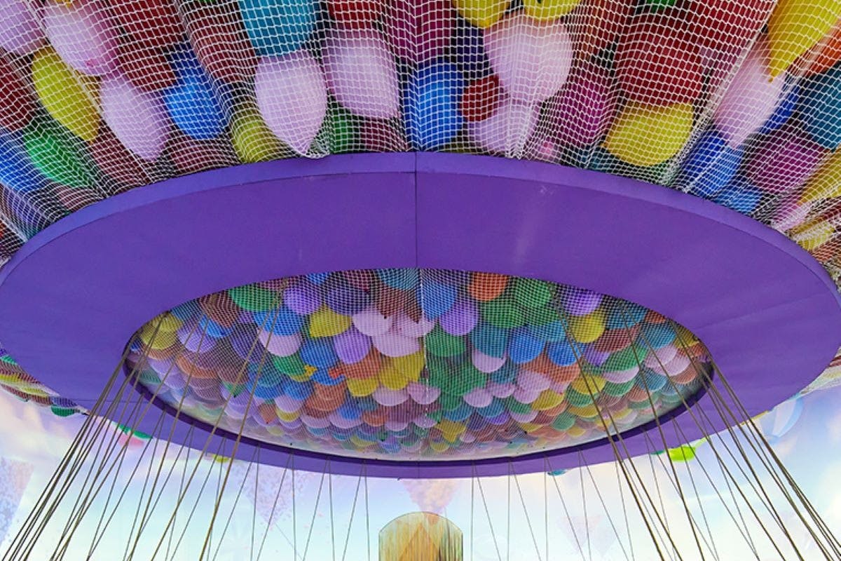 V síti, zavěšené na kruhové konstrukci s fialovým okrajem, je obsažen velký shluk barevných balónků.
