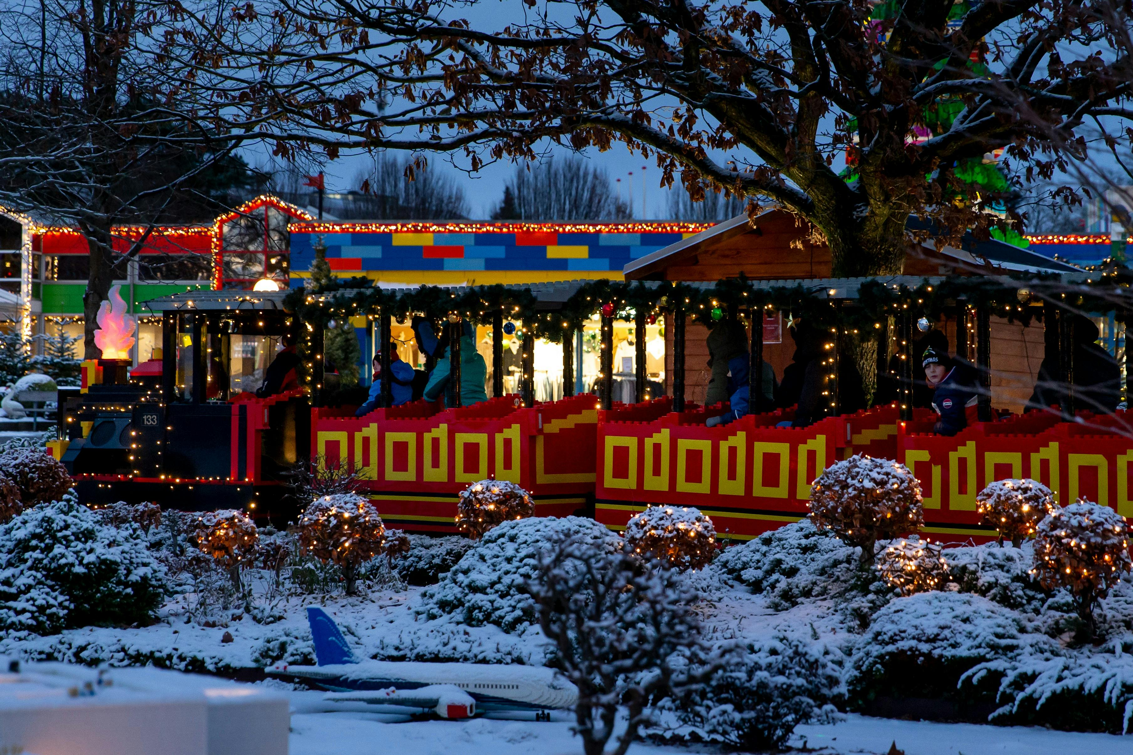 Праздничная поездка на поезде, украшенном огнями и гирляндами, проходит через снежную сцену с модельным самолетом неподалеку.