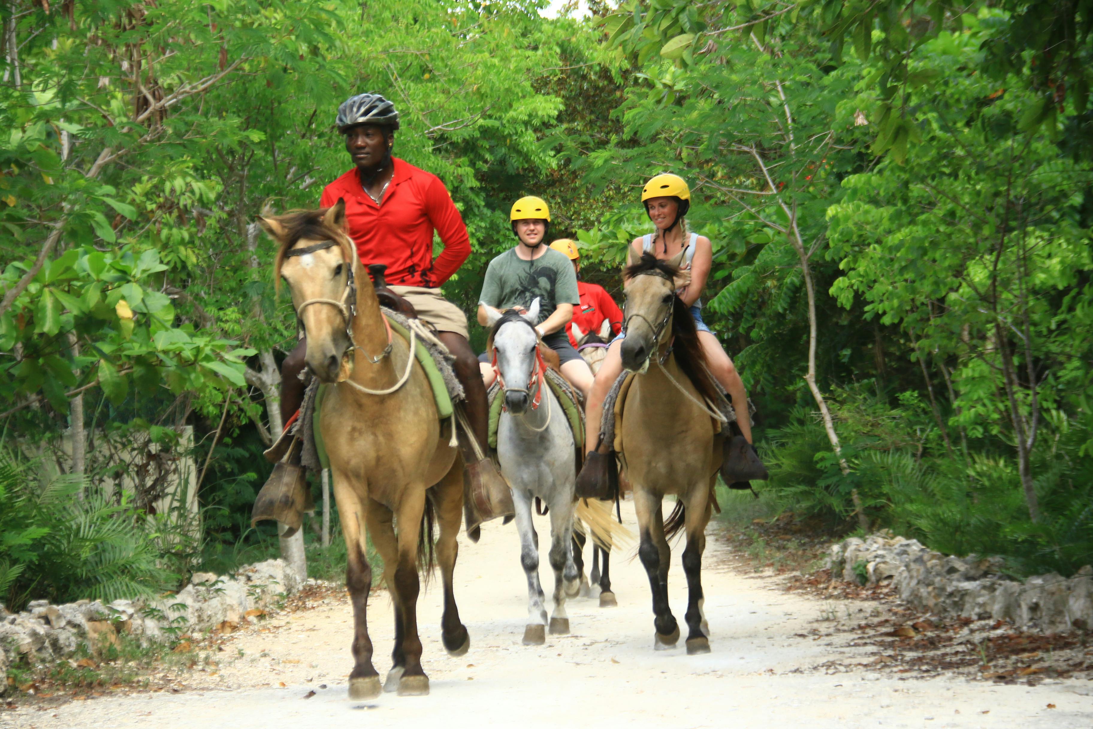 三个戴着头盔的人沿着森林小径骑马，在另一匹马上穿着红色衬衫的人的引导下。