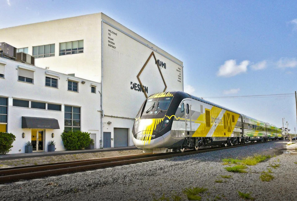 A modern Brightline train passing by a white building with "Miami Design District" signage on a clear day.