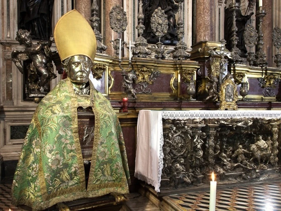 Gold statue of a bishop wearing a green and gold ornate robe and hat, situated beside a richly decorated altar.