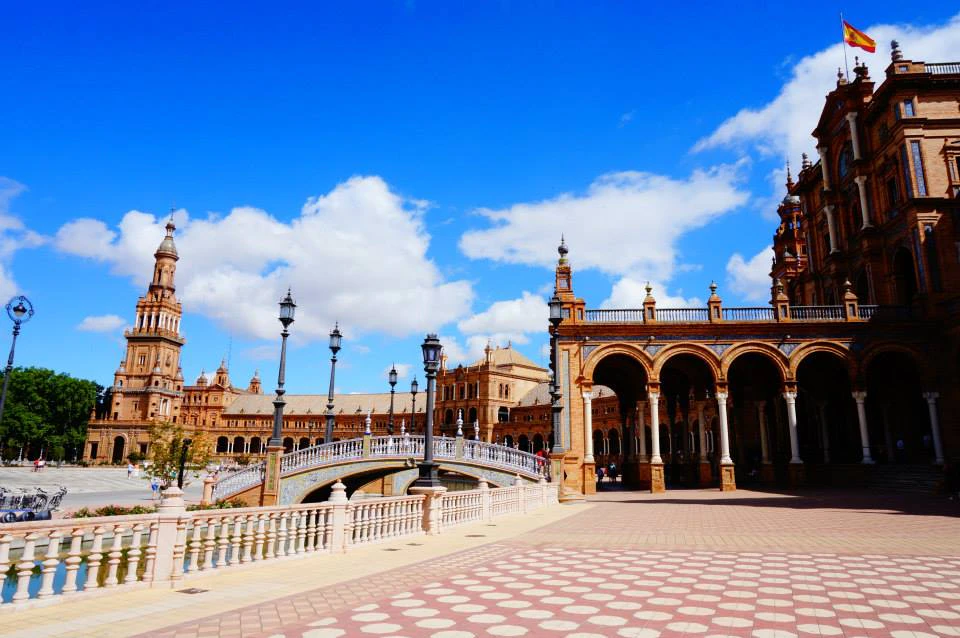 A plaza with arched architectural structures, ornate lampposts, and a bridge, under a bright blue sky with scattered clouds.