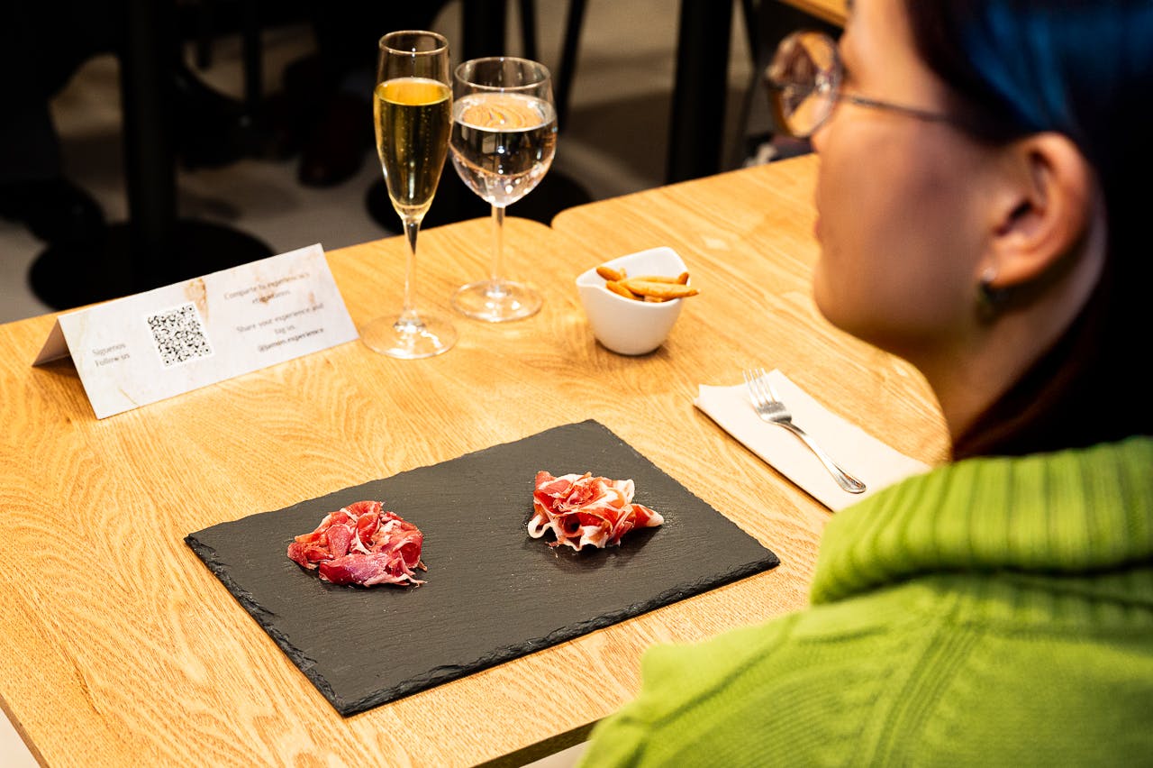Uma pessoa com uma camisola verde senta-se à mesa com um prato de ardósia com carne curada, copos de bebidas e uma tigela de pãezinhos.