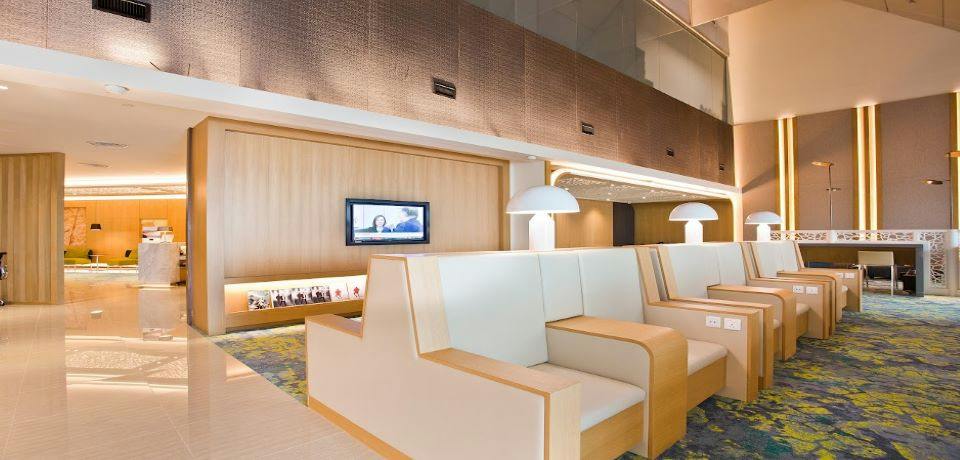 Modern lounge interior with beige seating, a wall-mounted TV, decorative lighting, and a wood-paneled wall.