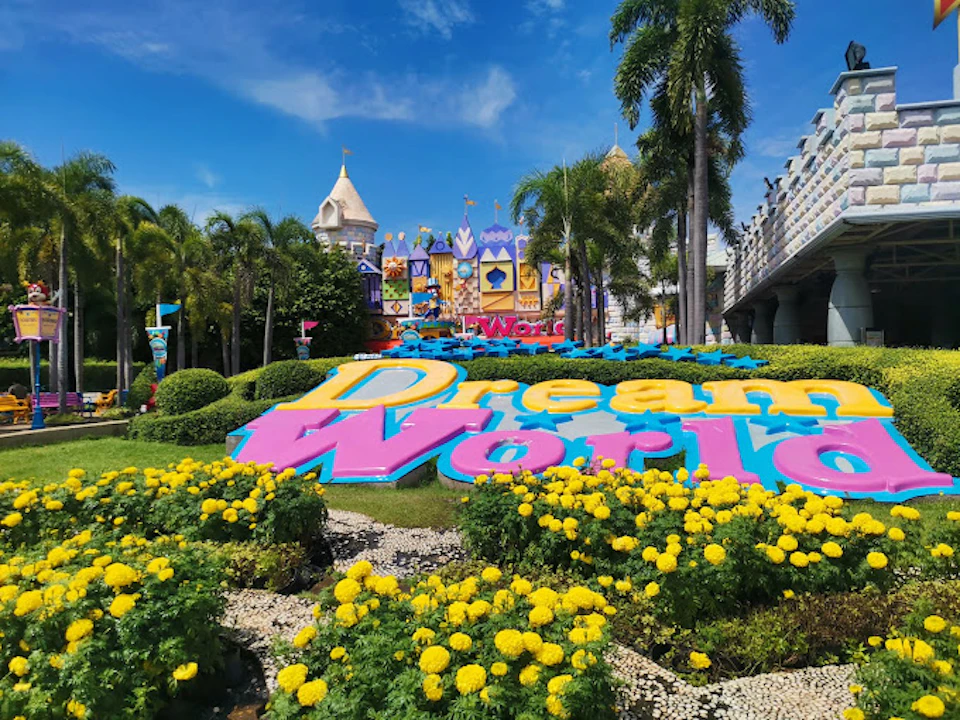 Colorful "Dream World" sign amidst vibrant flowers and lush greenery, with a castle-like building and palm trees in the background.