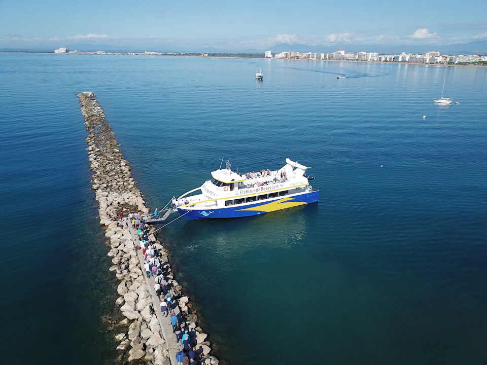 Aerial view of a boat docked at a rock pier, with a line of people waiting to board. The coastline and cityscape are visible in the distance.