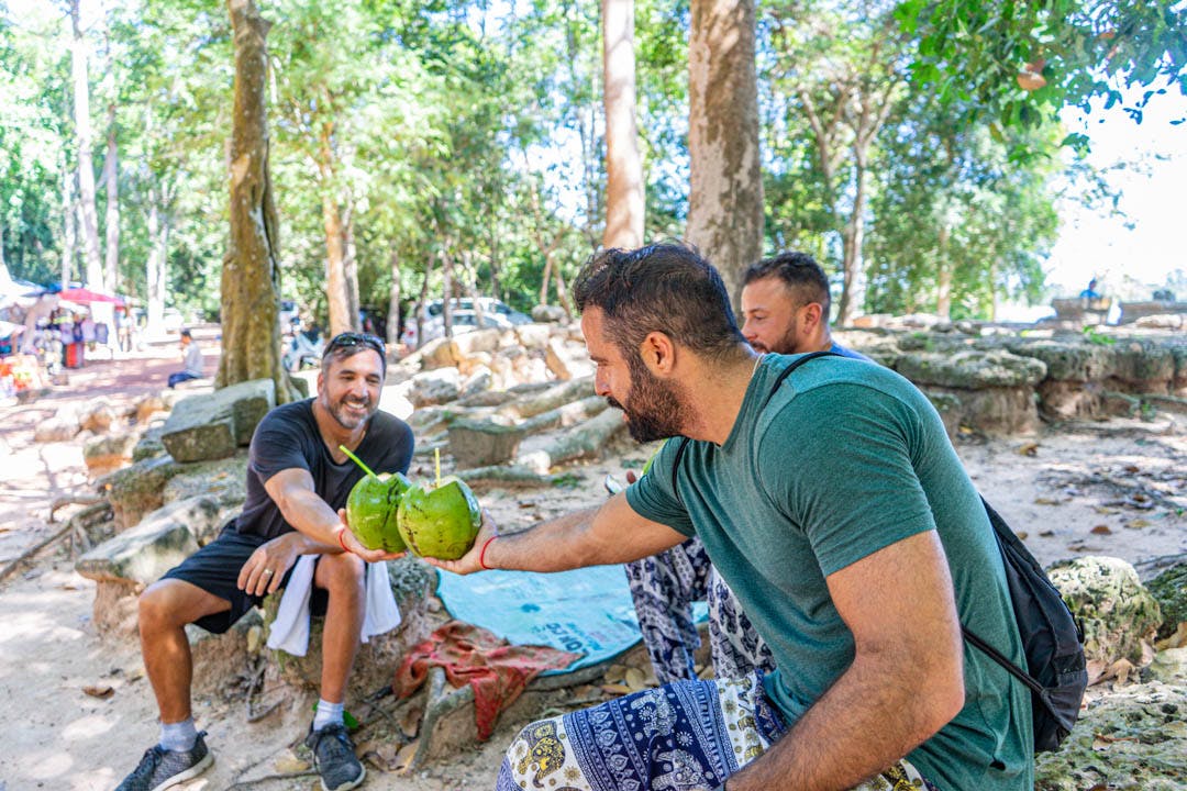 Tres hombres, sentados al aire libre con árboles y rocas alrededor, sonríen y entrechocan cocos con pajitas dentro.