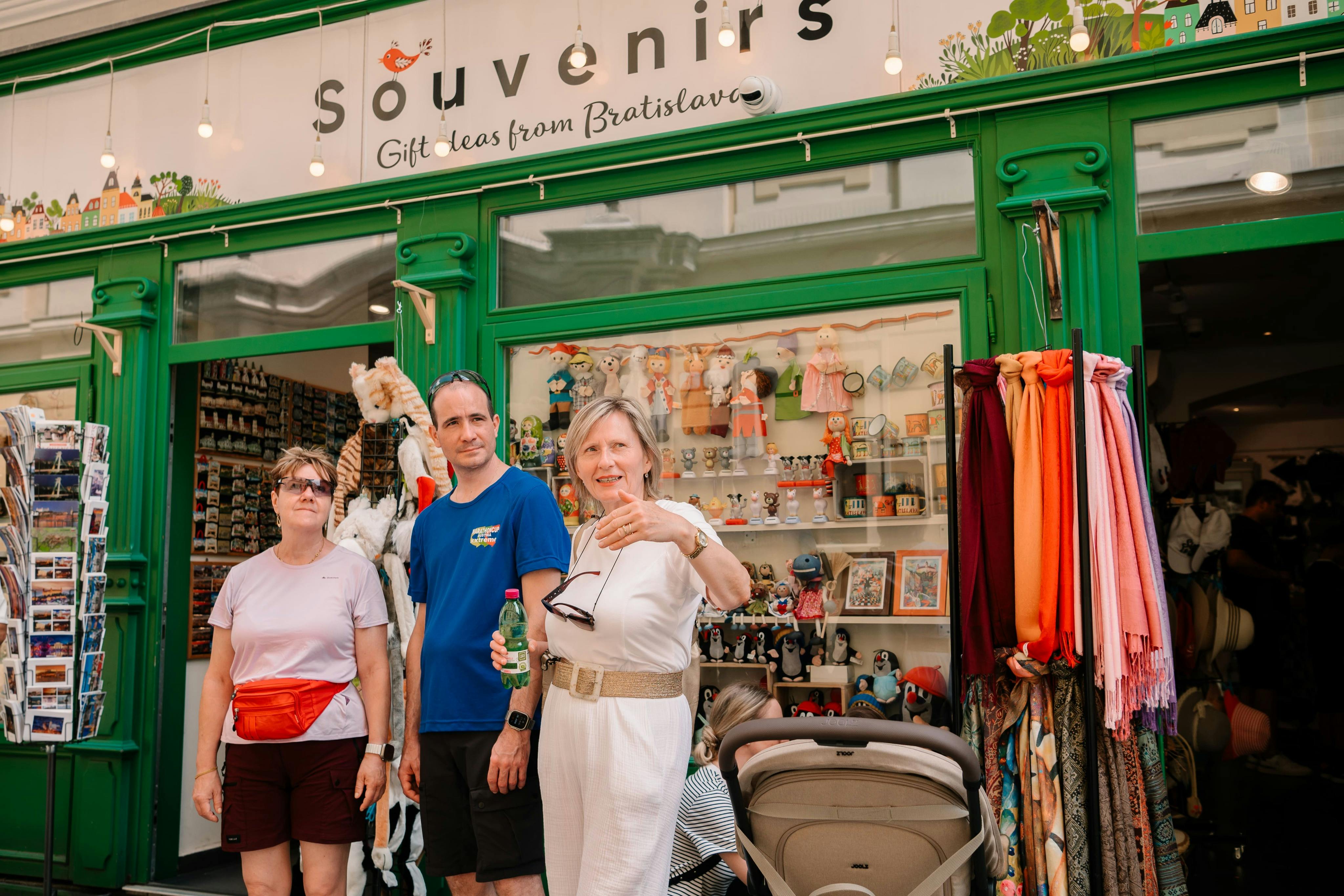 Τρία άτομα στέκονται μπροστά από ένα κατάστημα με σουβενίρ με πράσινη πρόσοψη. Η πινακίδα του καταστήματος αναφέρει: "Αναμνηστικά - Ιδέες για δώρα από την Μπρατισλάβα".