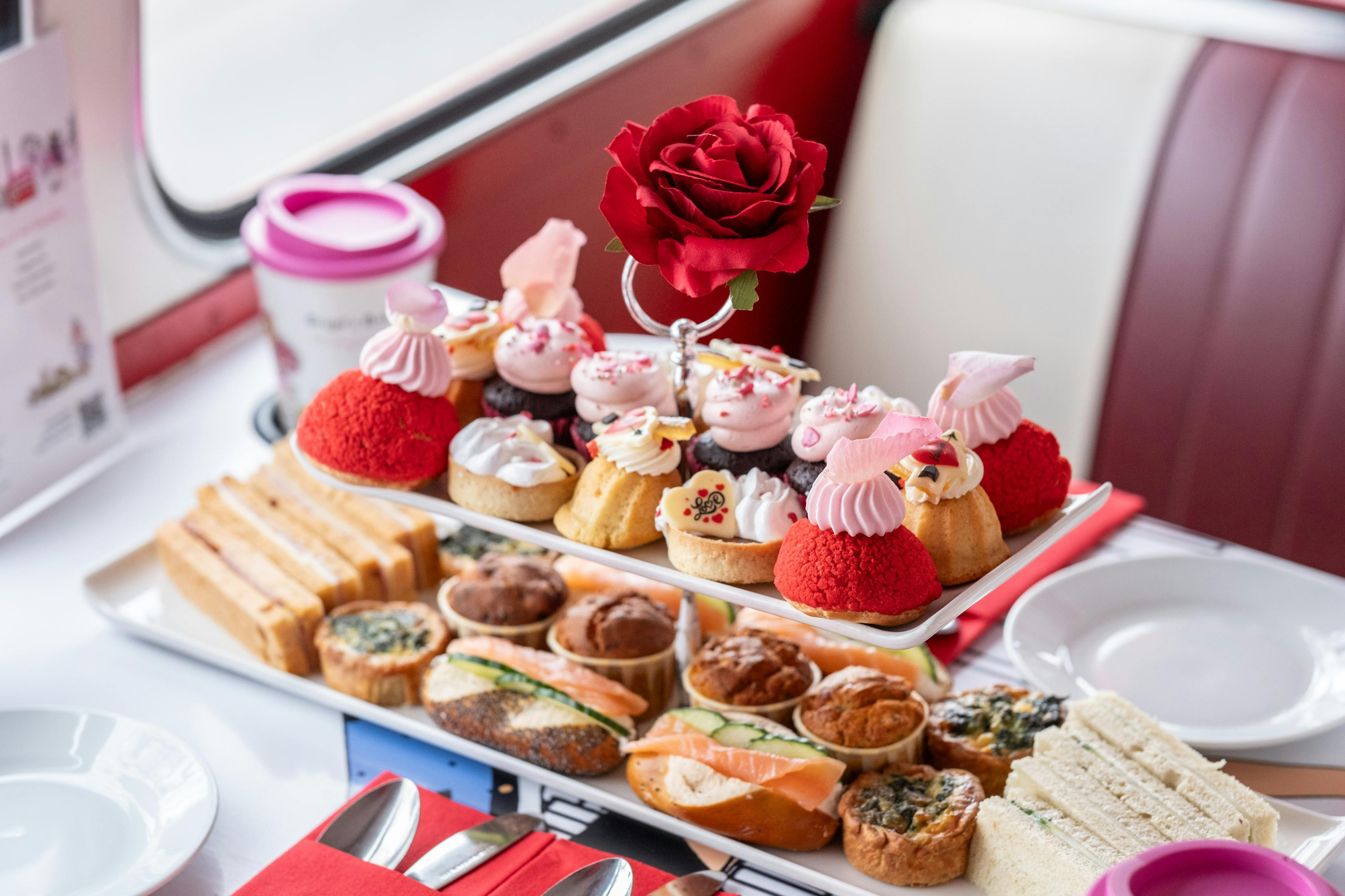 En todelt bakke fyldt med forskellige kager og sandwich, toppet med en rød rose, på et spisestel.