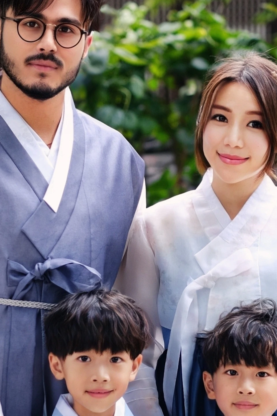 A family of four wearing traditional clothing poses outdoors near trees and a wooden door.