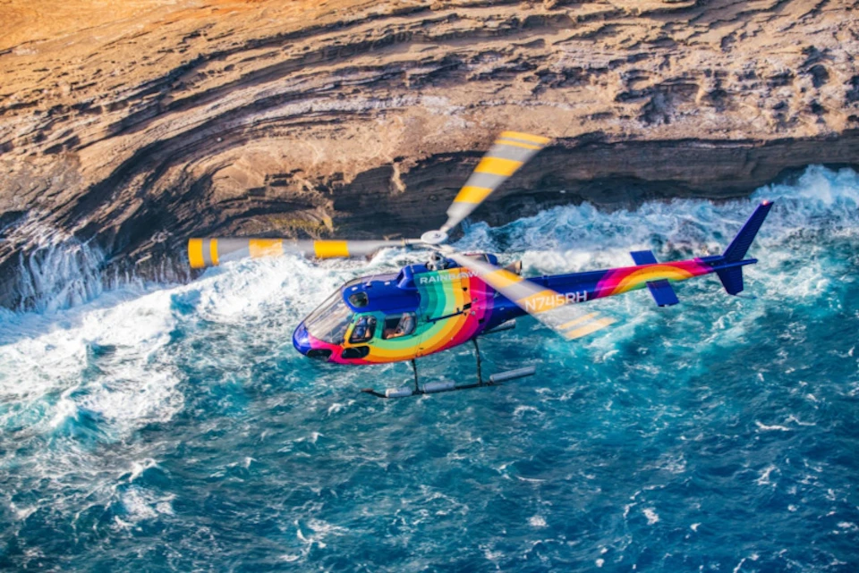 A colorful helicopter flies over blue ocean water near rocky cliffs.