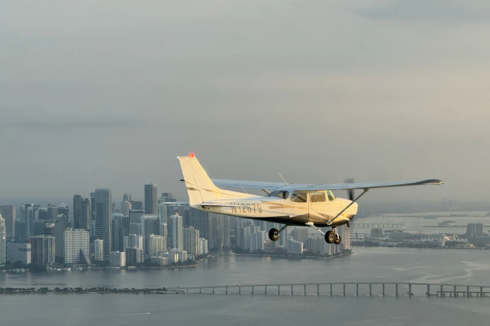 A small airplane flying over a cityscape with tall buildings and a bridge over the water below, under a cloudy sky.