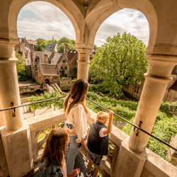 En kvinna och två barn har utsikt över en historisk murgröntäckt innergård från en stenbalkong med välvda pelare.