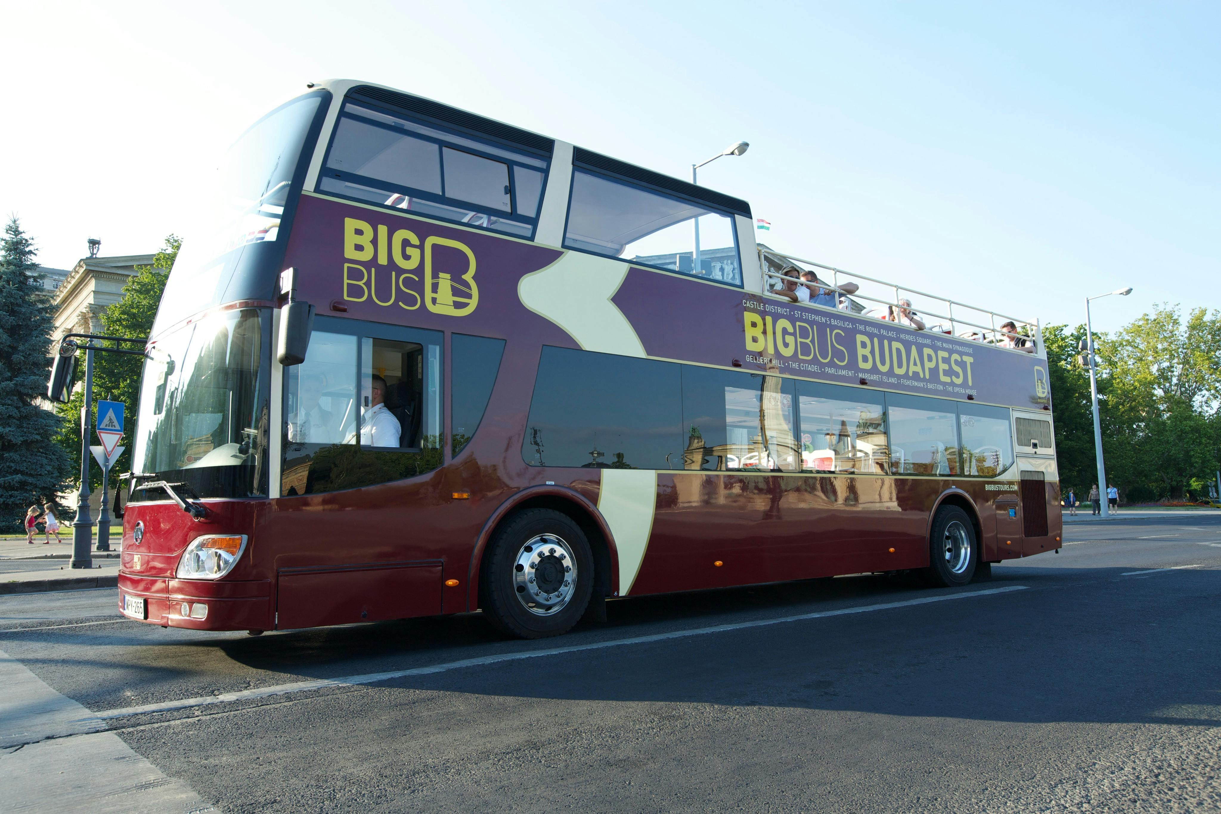 A maroon double-decker tour bus labeled "Big Bus Budapest" is parked on a road, with passengers seated on the upper deck.