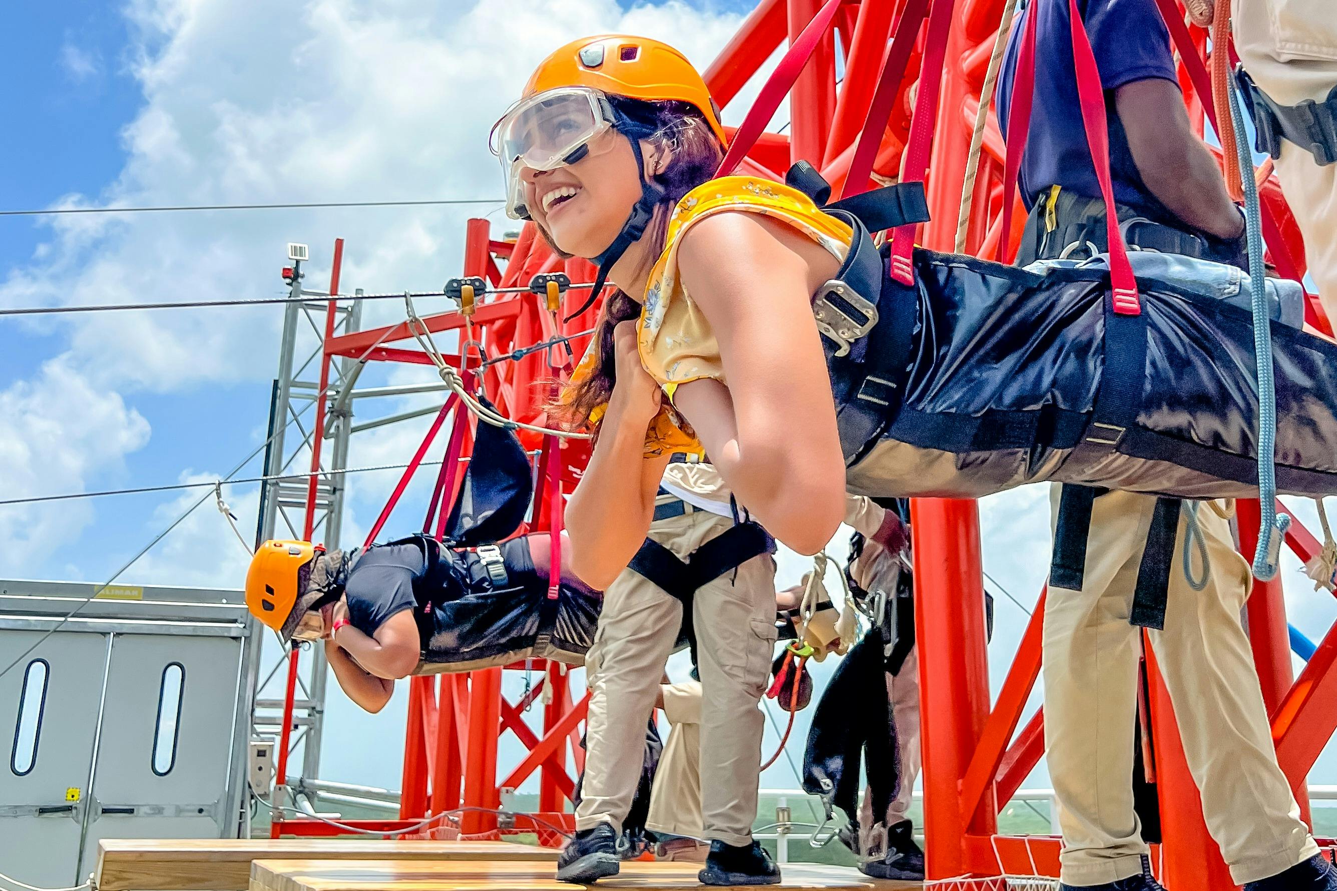 People wearing harnesses and helmets prepare to zipline from a tall platform with red support structures under a blue sky.