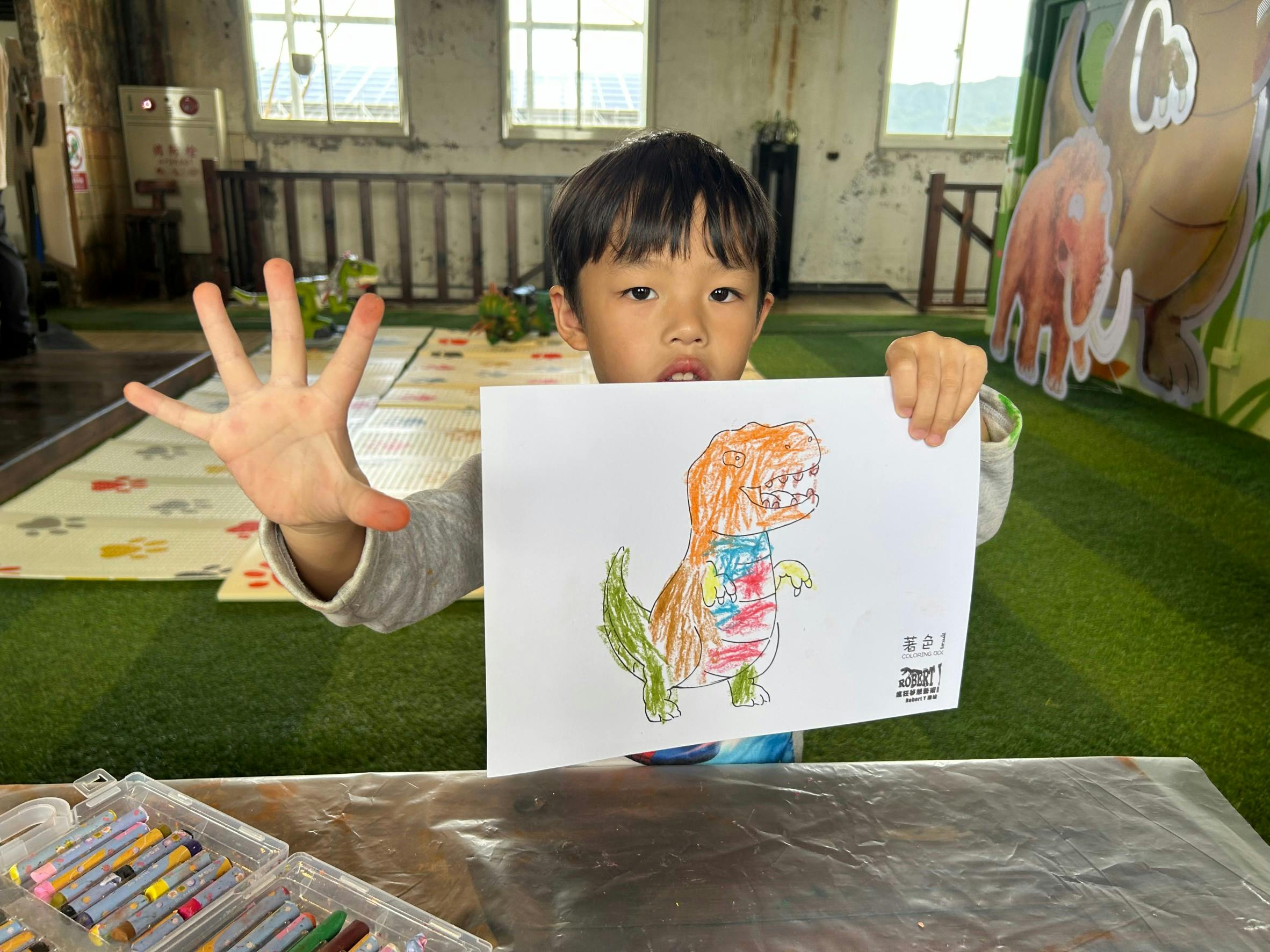A child holds up a drawing of a colorful dinosaur in a room with green flooring and large windows.