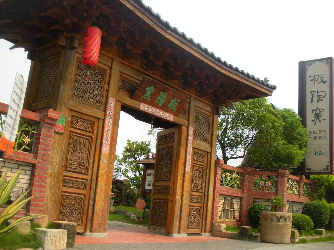 A traditional wooden gate with intricate carvings and a red lantern, surrounded by brick and greenery.