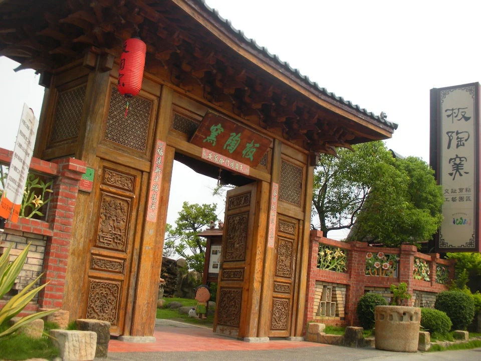 A traditional wooden gate with intricate carvings and a red lantern, surrounded by brick and greenery.