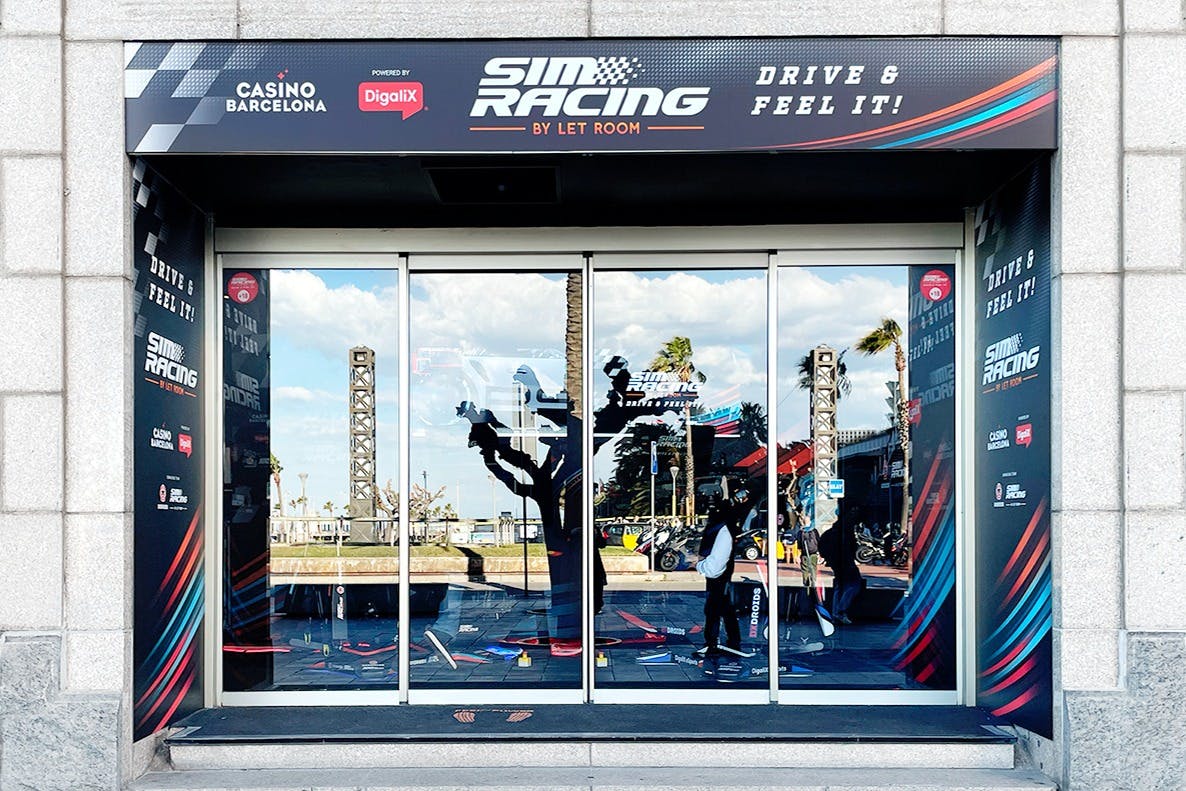 Widoczne szklane drzwi „Sim Racing By Let Room” z odbiciami drzew i konstrukcji, sztandarami promocyjnymi i kilkoma osobami.