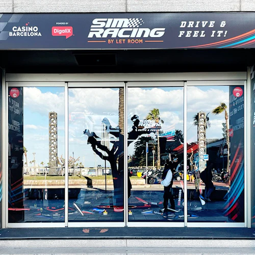 Glasdörrar av "Sim Racing By Let Room" med reflektioner av träd och strukturer, PR-banderoller och några personer synliga.