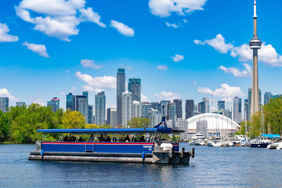 Boat on water with skyline of tall buildings in the background under a blue sky with clouds. Trees and yachts are also visible.