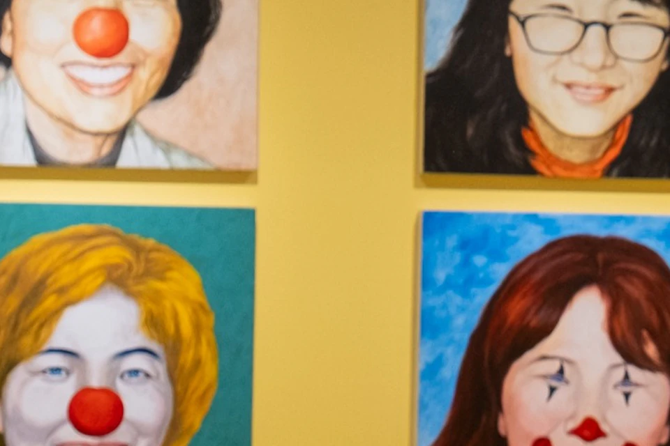 An art exhibit featuring colorful portraits on yellow walls, hanging white sculptures, and display pedestals with books.
