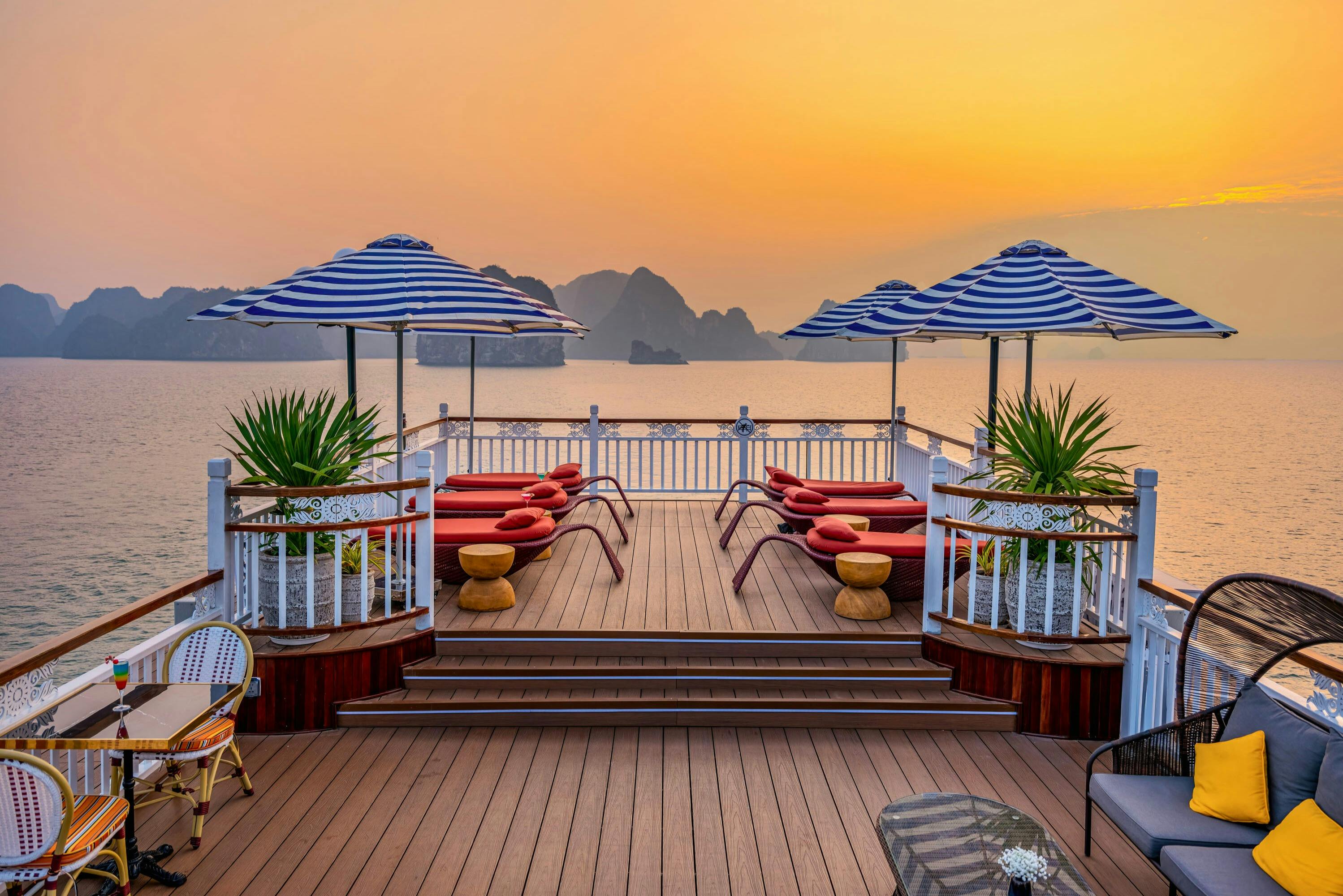 Deck with striped umbrellas, lounge chairs, and small tables overlooking a body of water and rocky islands at sunset.