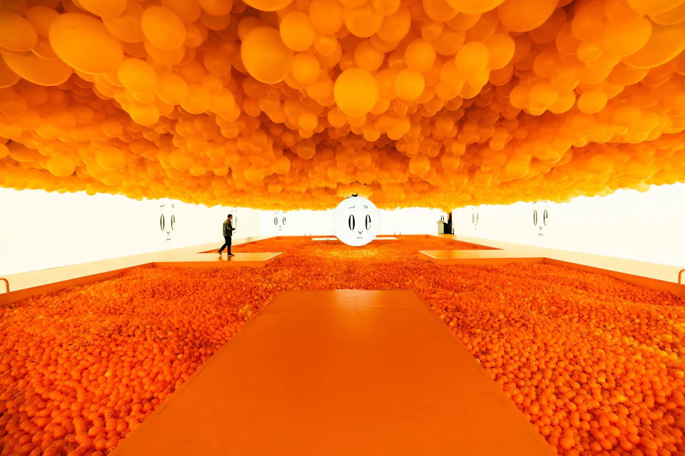 Room filled with orange balloons on ceiling, orange ball pit on floor, and a person walking near a white sphere with eyes.