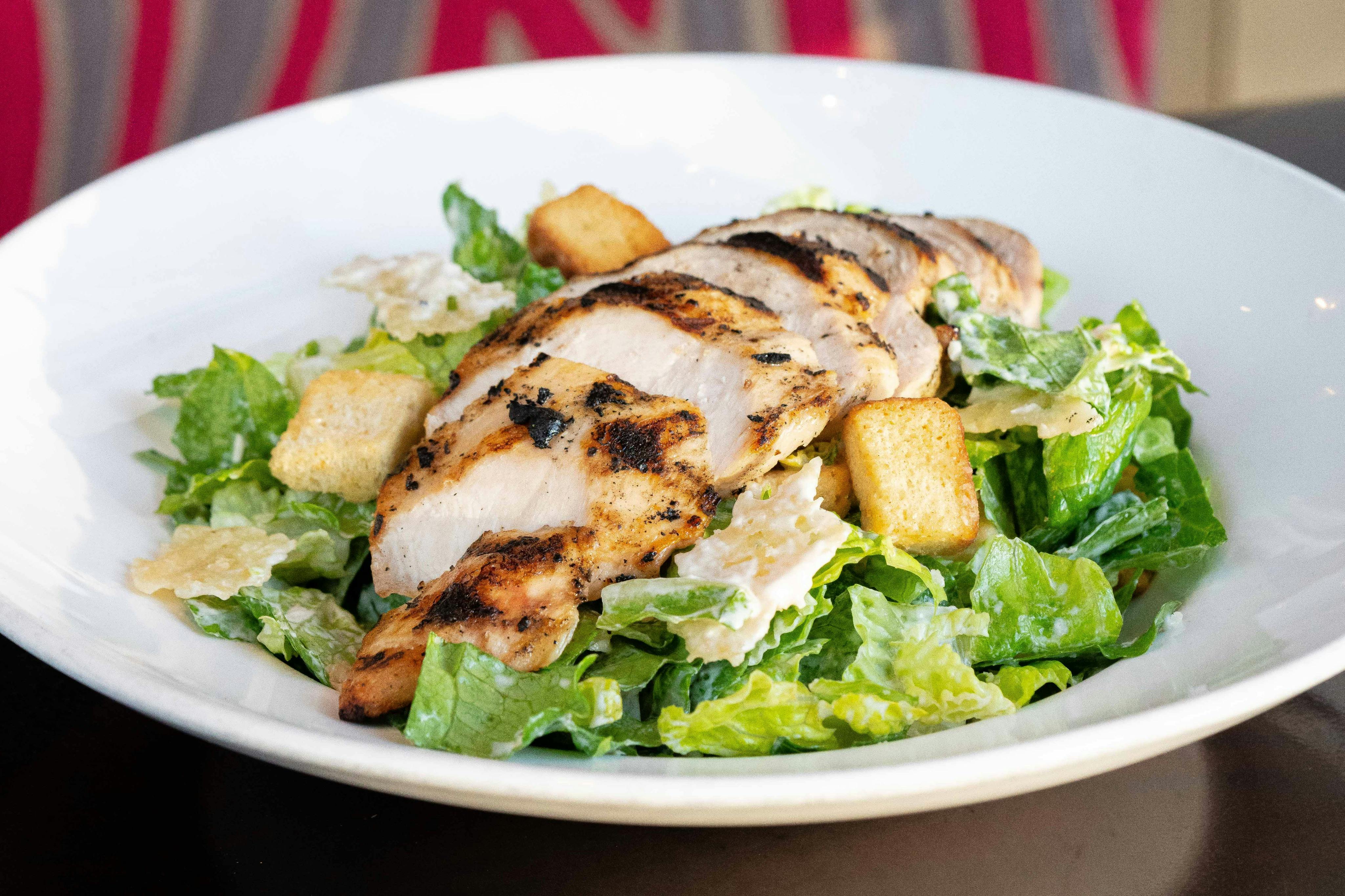 A Caesar salad topped with sliced grilled chicken breast and croutons in a white bowl.