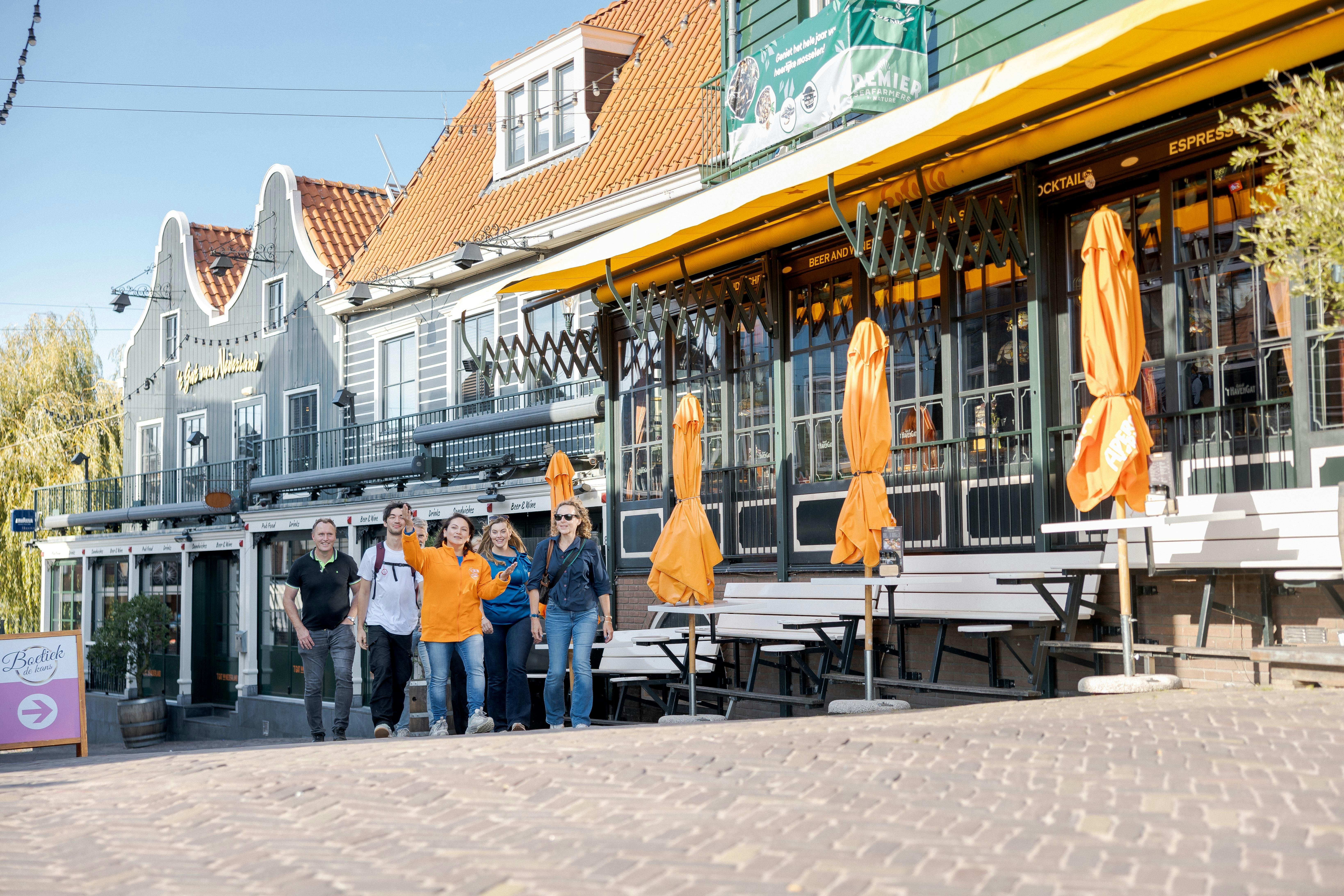 A group of five people walks along a cobblestone street with colourful buildings and outdoor seating with orange umbrellas in the background.
