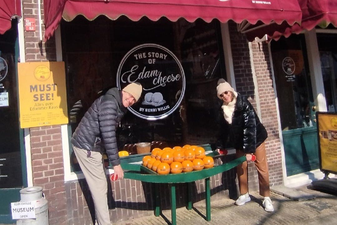 Δύο άνθρωποι με χειμερινά ρούχα στέκονται δίπλα σε μια βιτρίνα με τροχούς τυριού έξω από ένα κατάστημα με μια πινακίδα που γράφει «Η ιστορία του τυριού Edam».