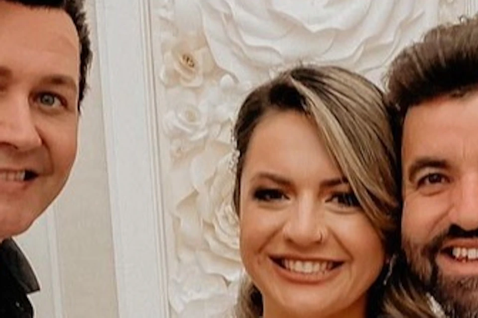 A couple, with the bride in a lace wedding dress holding a bouquet and the groom in a grey suit beside them, poses with a man in a gold suit jacket giving a thumbs up.