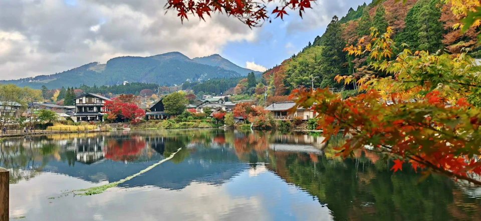 A scenic view of a tranquil lake reflecting autumn trees, traditional houses, and mountains under a cloudy sky.