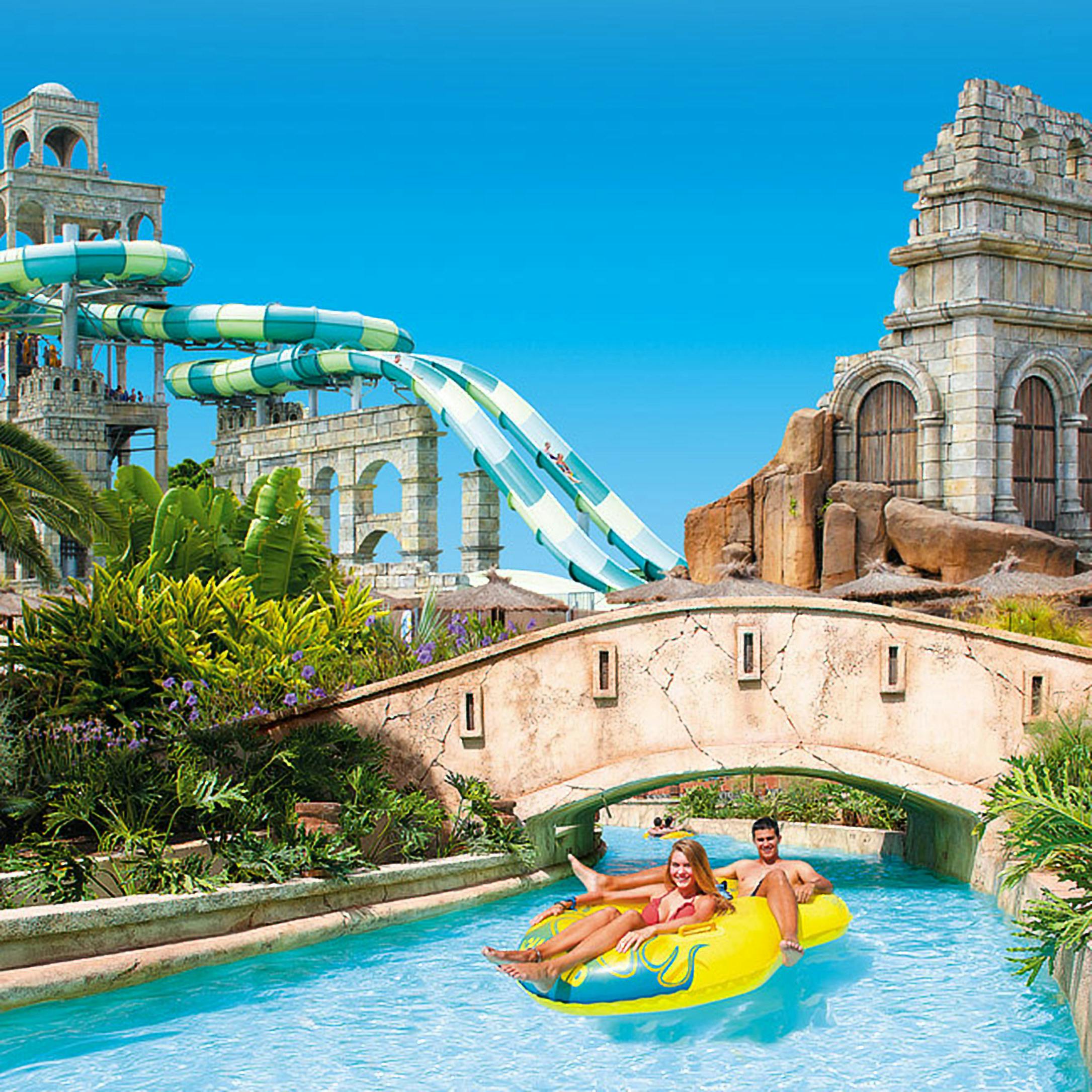 Two people float on yellow inflatable tubes in a lazy river with water slides and ancient ruins in the background.