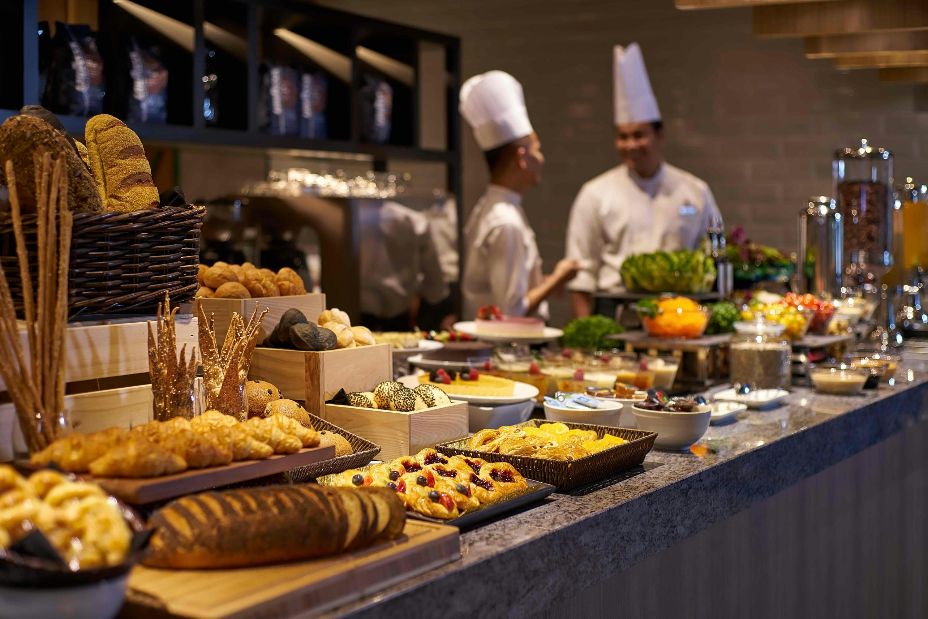 A buffet setup with various food items displayed on a counter. Two chefs in white uniforms are conversing in the background.