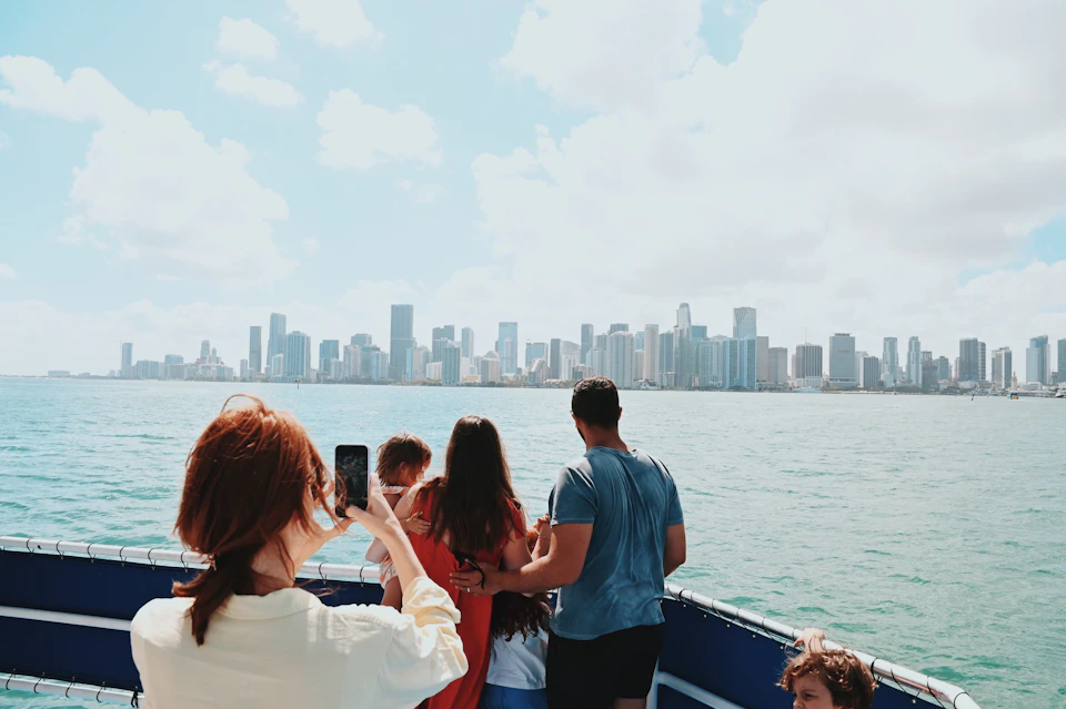People on a boat looking towards a city skyline across the water, with one person taking a photo using a smartphone.