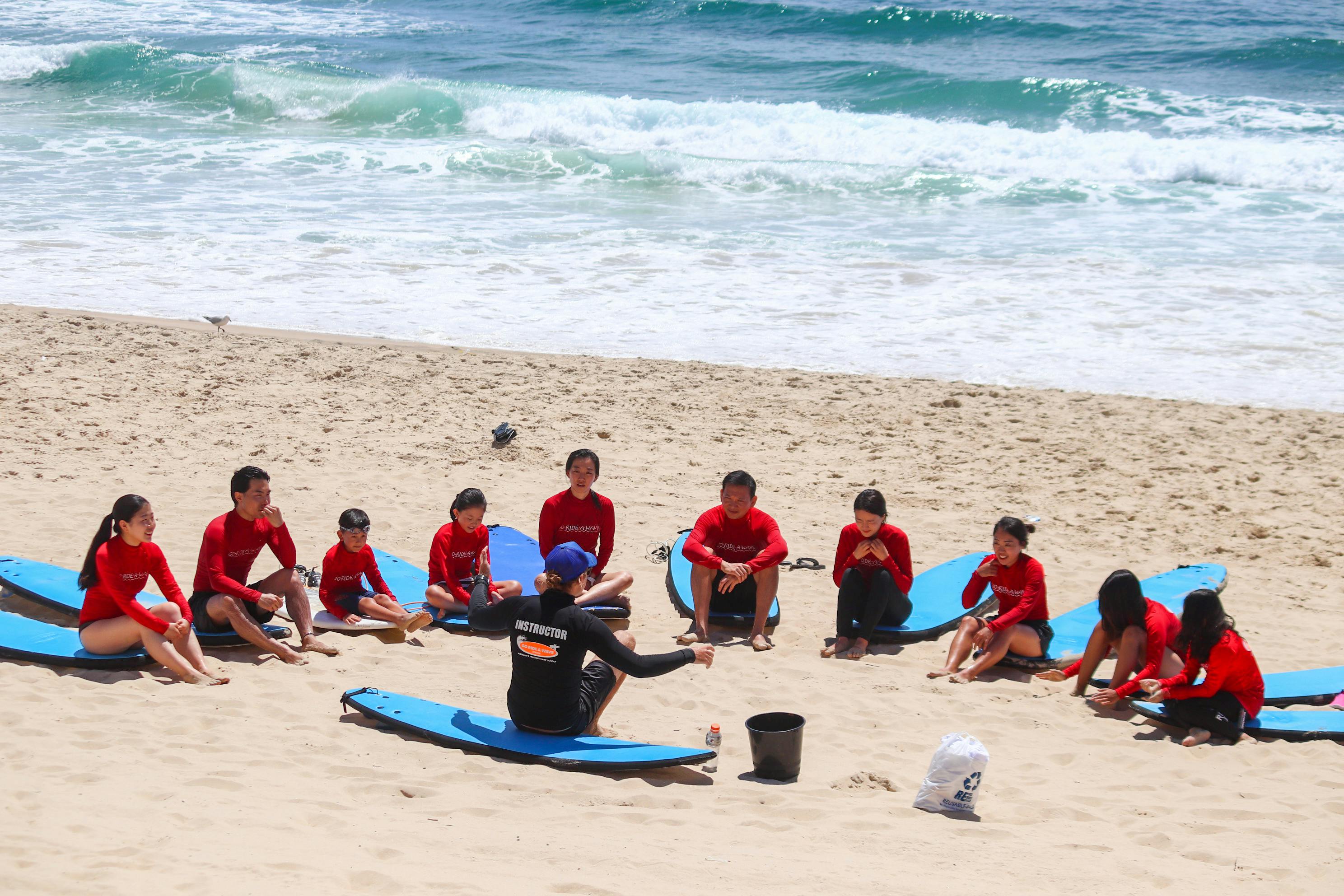 서핑보드가 달린 빨간 셔츠를 입은 한 무리의 사람들이 바다 근처의 모래 해변에 앉아 강사의 말을 듣고 있습니다.