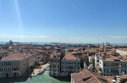 Vista sopraelevata di una città con tetti di tegole rosse, canali, barche ed edifici storici sotto un cielo azzurro e limpido.