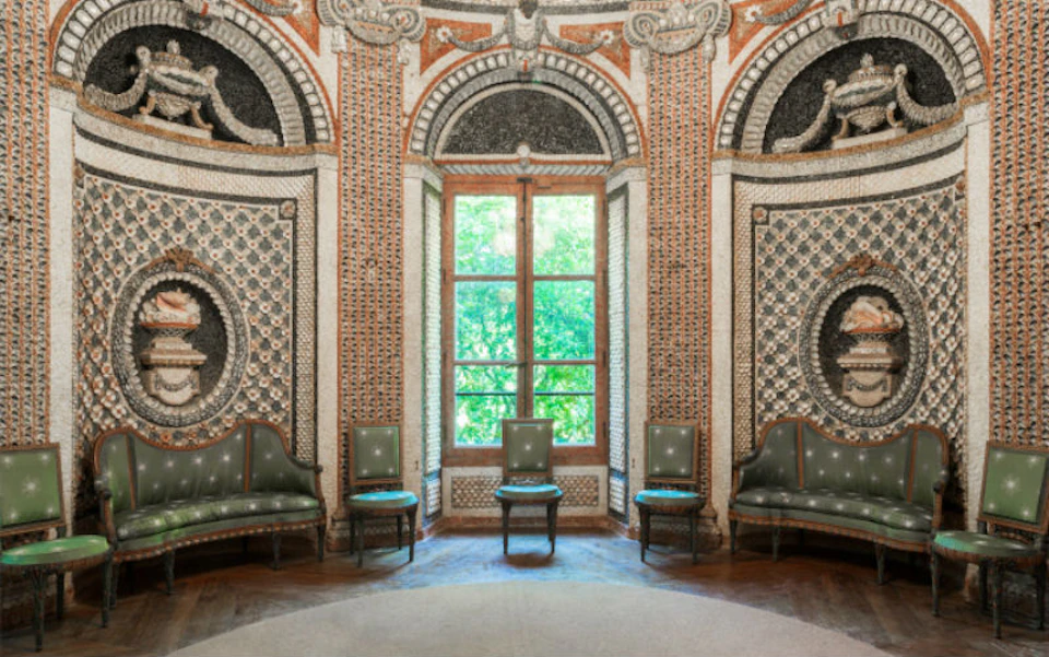 An ornate room with intricate wall designs, four green chairs, and a large window showcasing lush greenery outside.