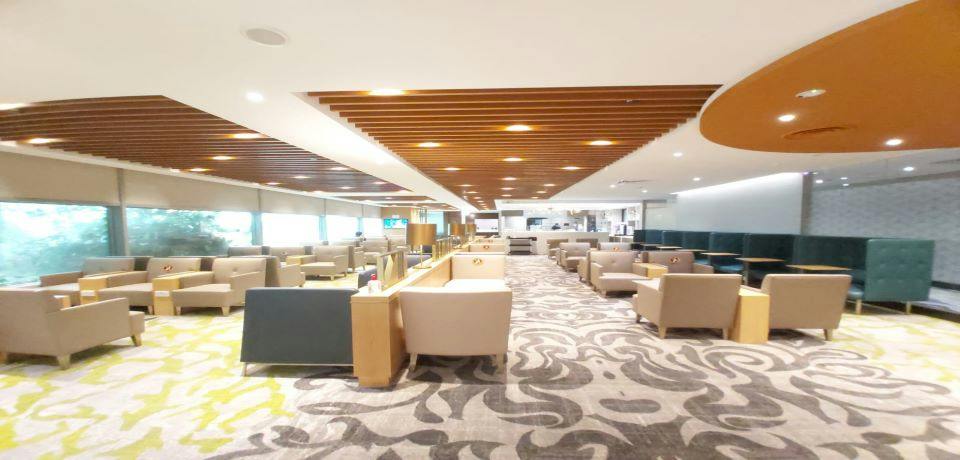 A spacious lounge with armchairs, tables, and a patterned carpet, featuring a wooden slat ceiling and ambient lighting.