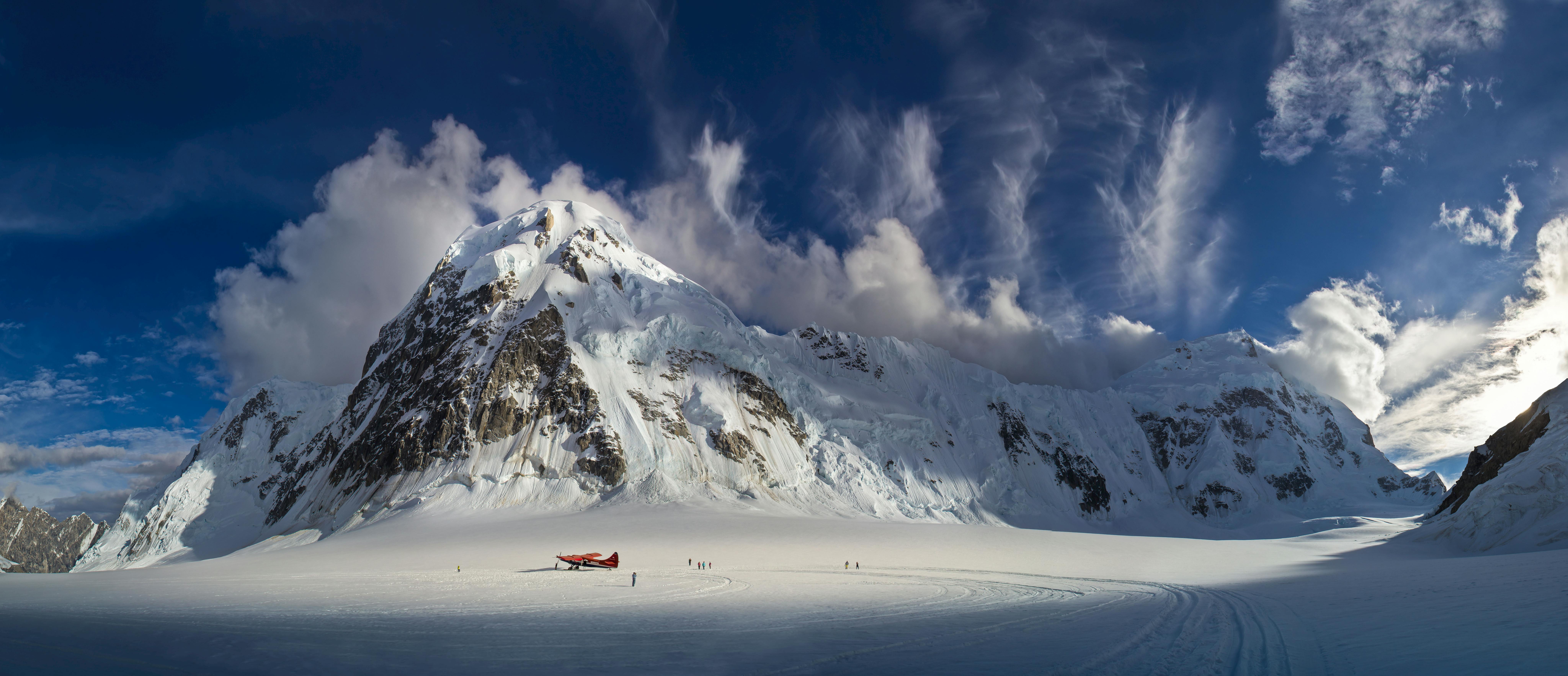 Un avion rouge est posé sur une plaine enneigée, au pied d'une imposante montagne enneigée, avec des nuages épars et un ciel bleu clair au-dessus.