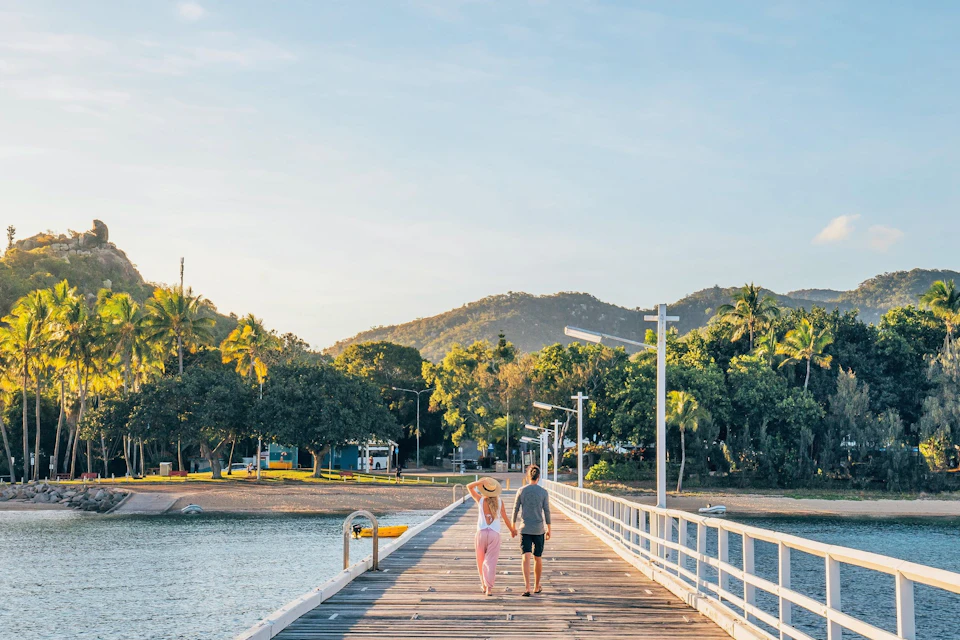 A couple walks hand in hand on a wooden pier surrounded by water, with trees, hills, and a clear sky in the background.