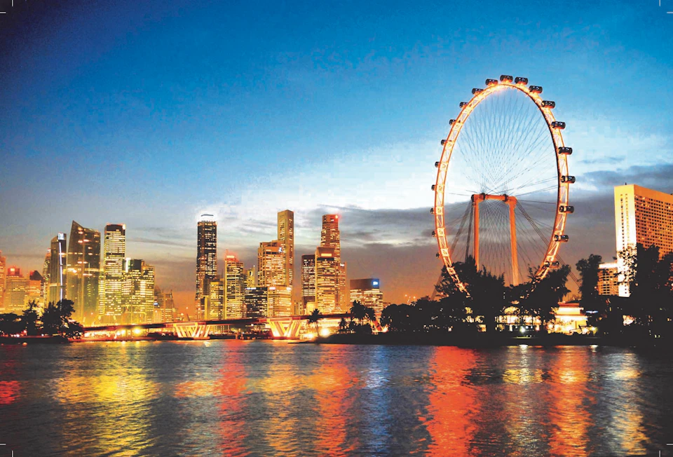 City skyline at dusk with illuminated skyscrapers and a large Ferris wheel by the water, reflecting colorful lights.