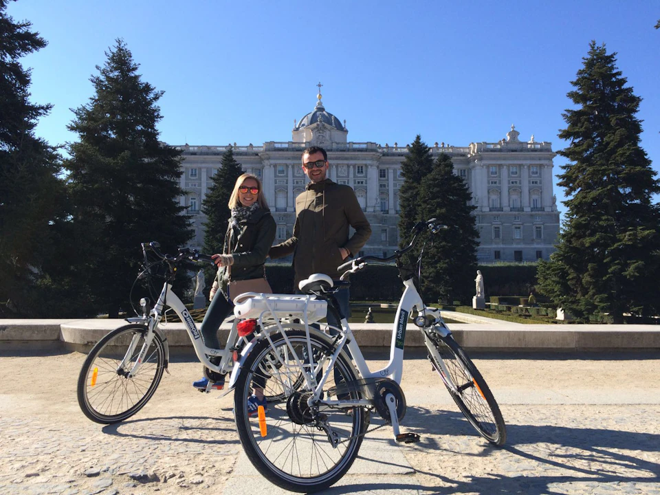 Two people stand with bicycles in front of a large historic building, surrounded by trees and a clear, sunny sky.
