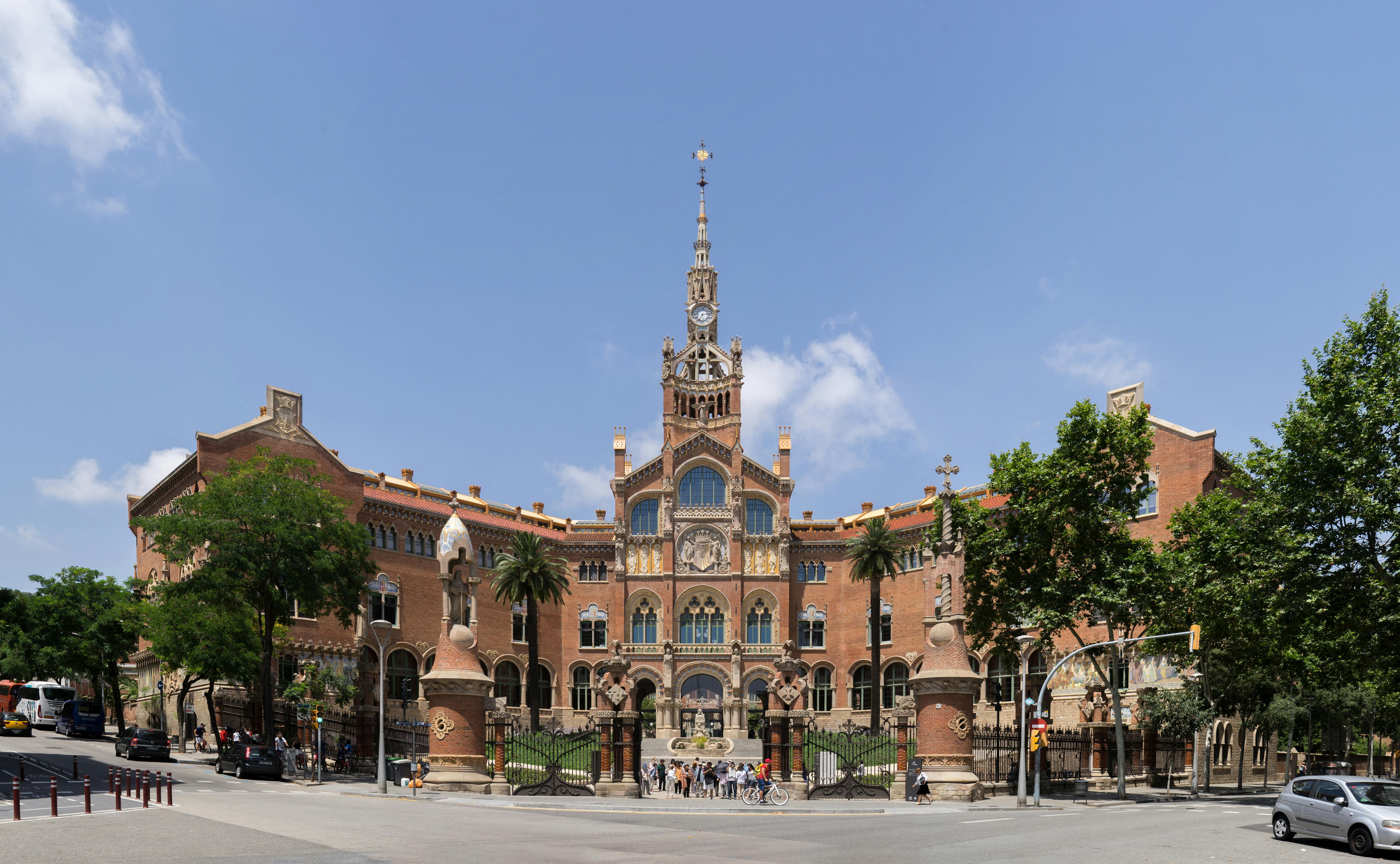 People stand in front of a large, ornate, red-brick building with a central tower, under a clear blue sky.