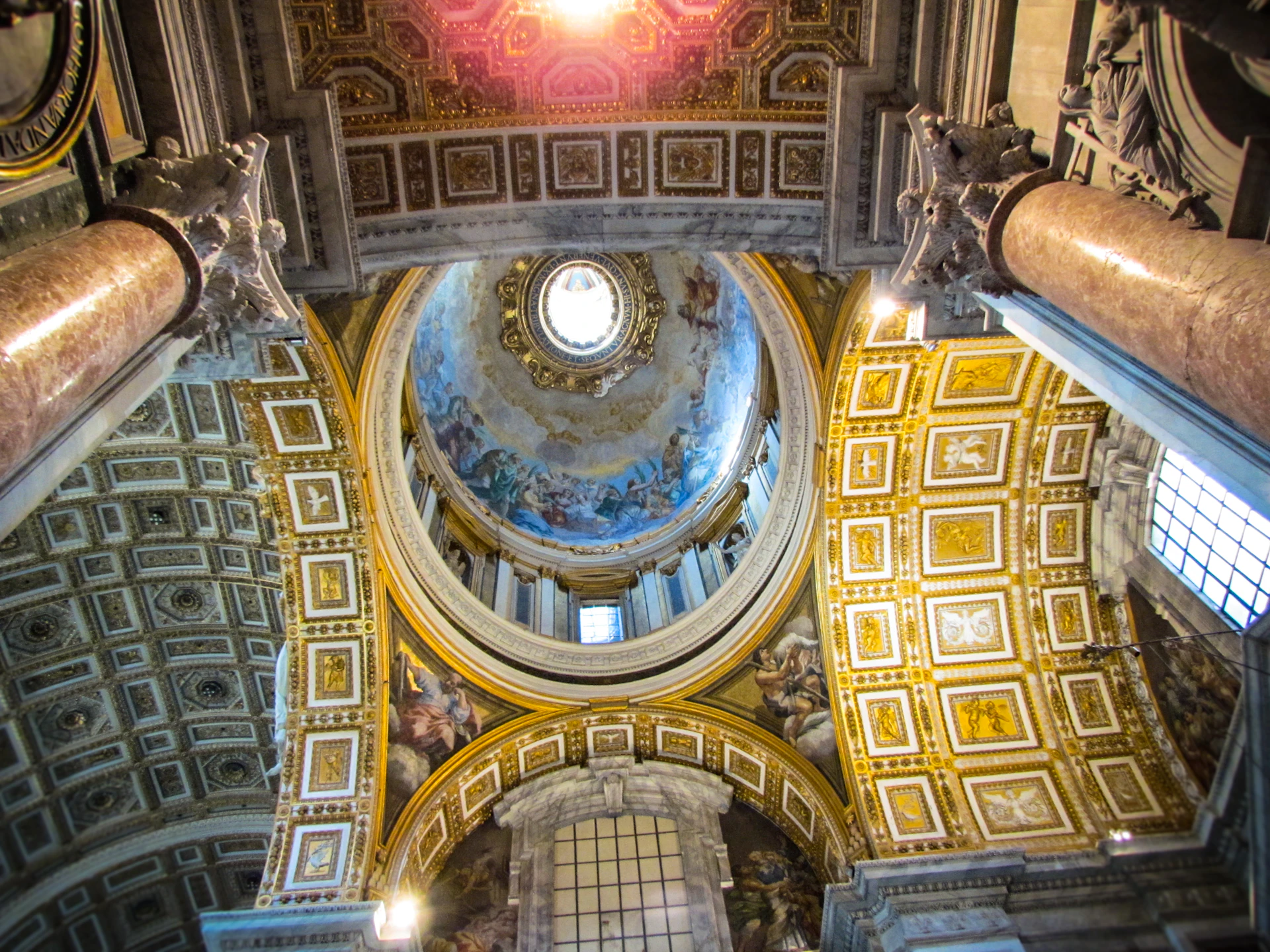 Ornate, gold-trimmed church ceiling with detailed frescoes, a central dome, and decorative pillars. Bright light shines from the dome.
