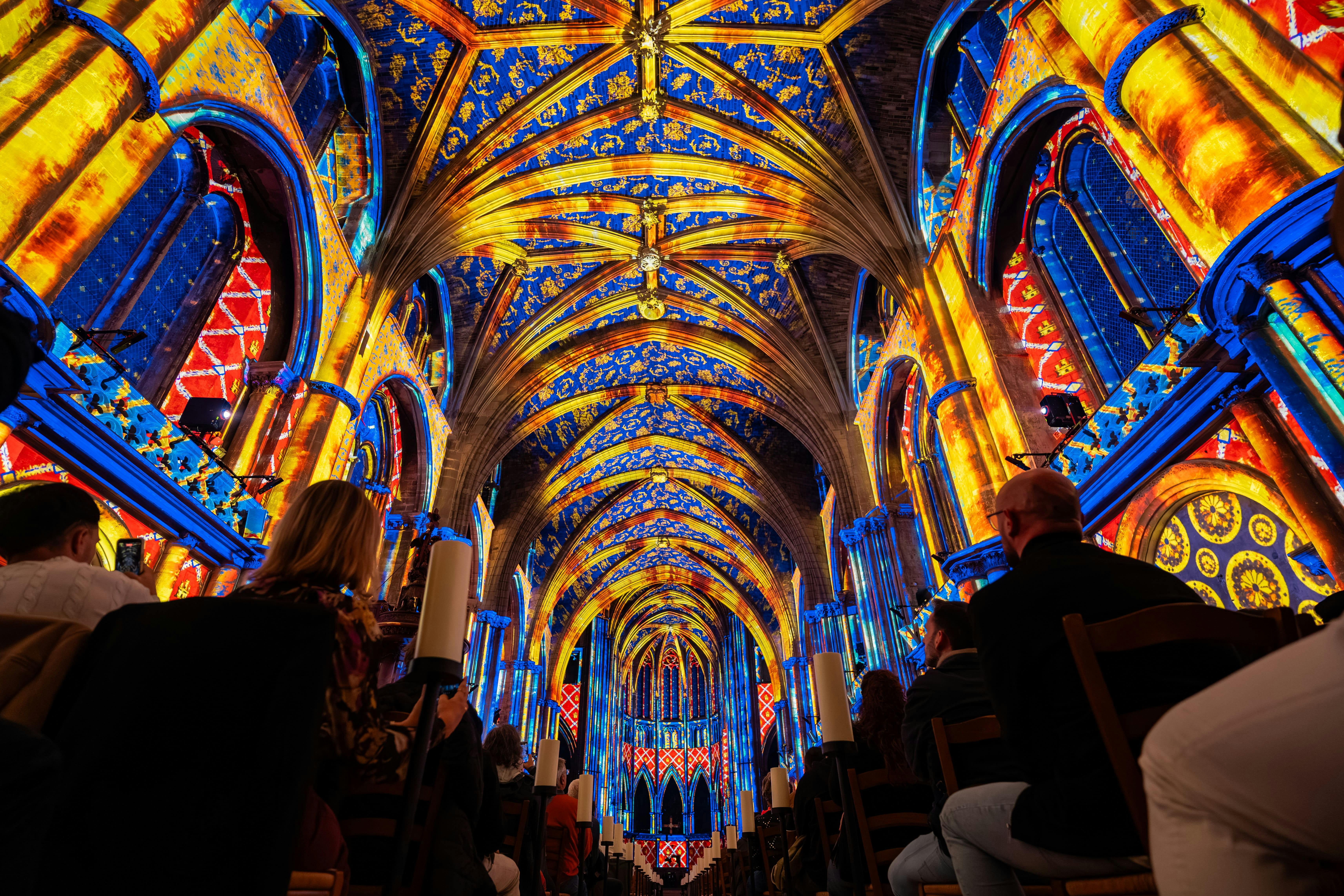 People seated in a cathedral with brightly illuminated, colorful vaulted ceilings and arches displaying intricate light patterns.